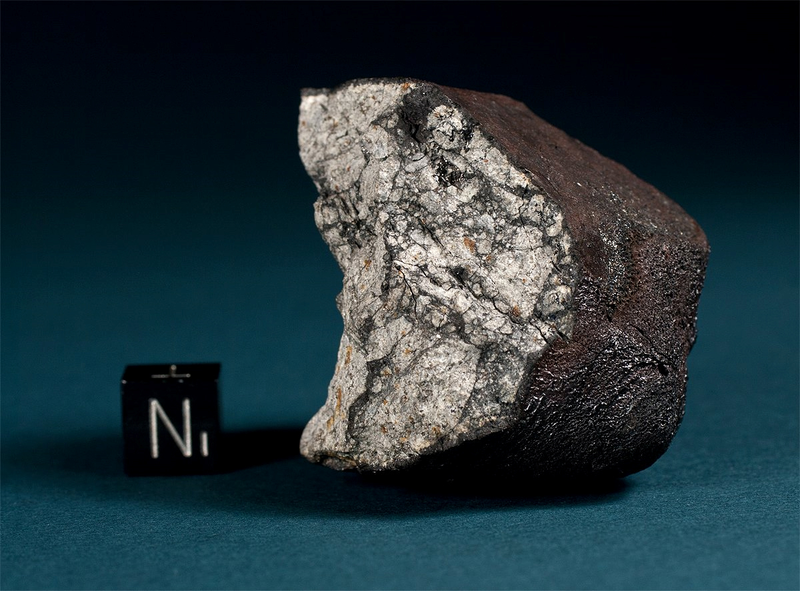 A 3.96 oz Chelyabinsk meteorite chunk next to a 1 cm cube. The broken fragment displays a thick primary fusion crust with flow lines and a heavily shocked matrix with melt veins and planar fractures.